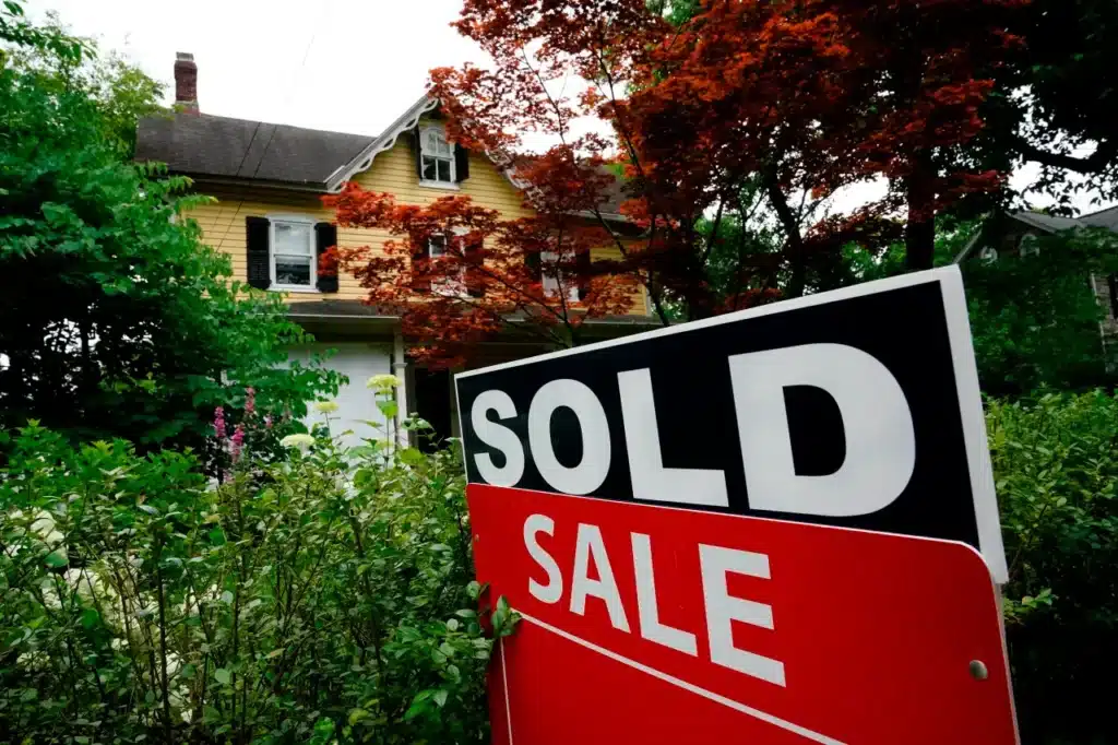 A prominent "SOLD" sign in black and white lettering placed over a red "FOR SALE" sign, situated in a lush garden with a yellow two-story house in the background.