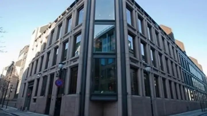 A low-angle shot of a multi-story modern office building on a street corner, featuring a light brown stone facade and large glass windows.