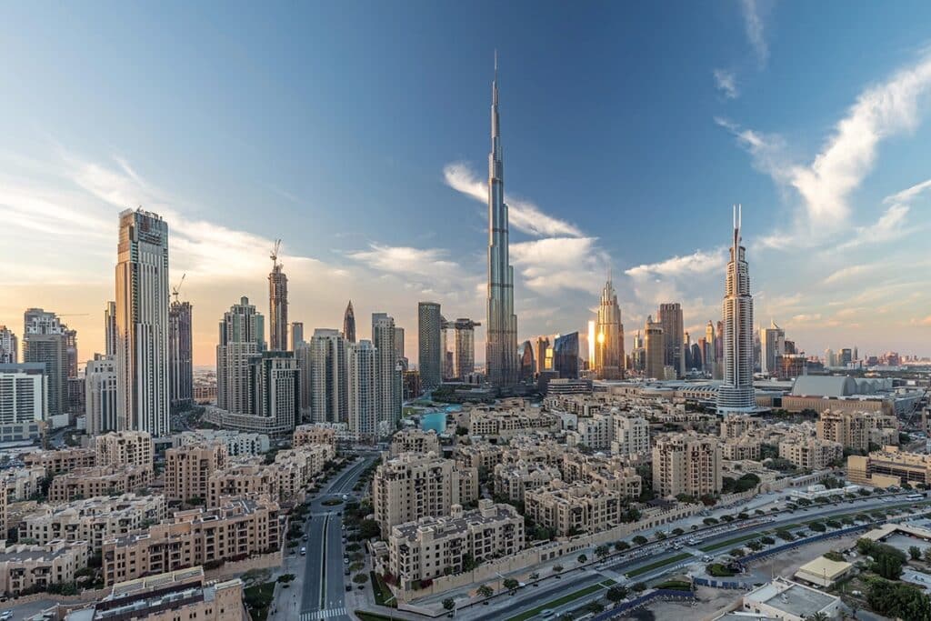A wide panoramic view of the Downtown Dubai skyline featuring the Burj Khalifa towering over surrounding skyscrapers and residential buildings under a bright blue sky with wispy clouds.