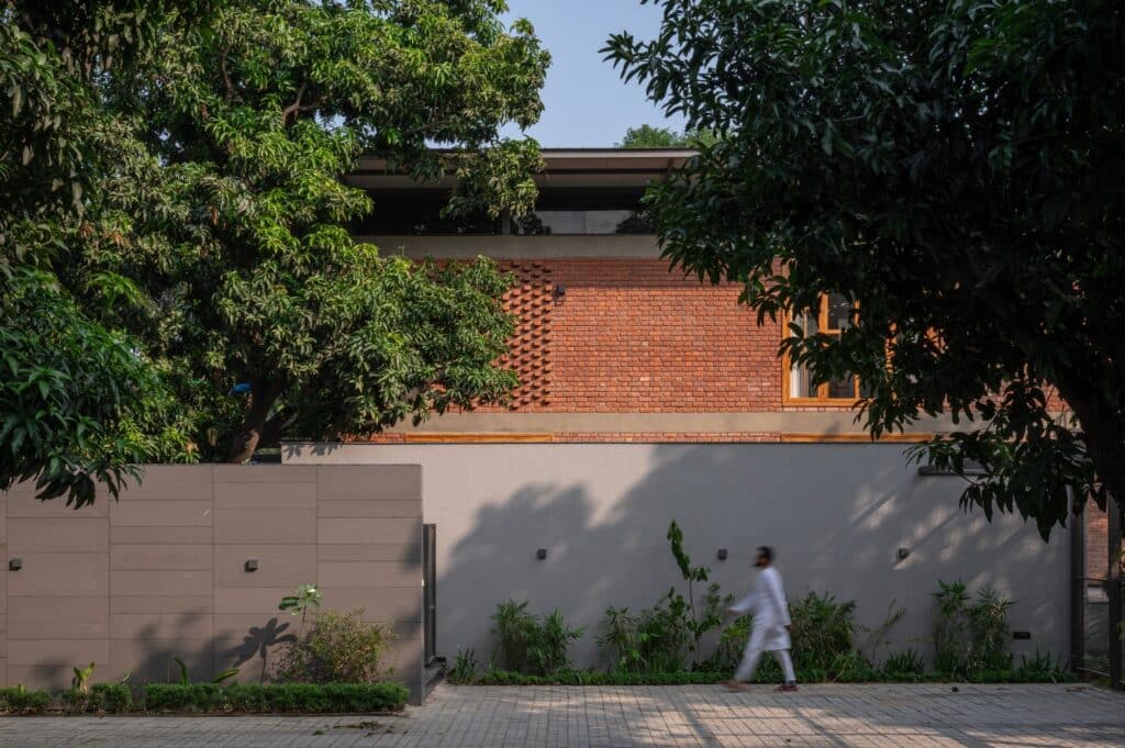 A detailed architectural view of the Gorakhpur Farmhouse exterior, focusing on the textured red brick walls, the upper terrace with a metal railing, and the integration of mature trees into the building's footprint.