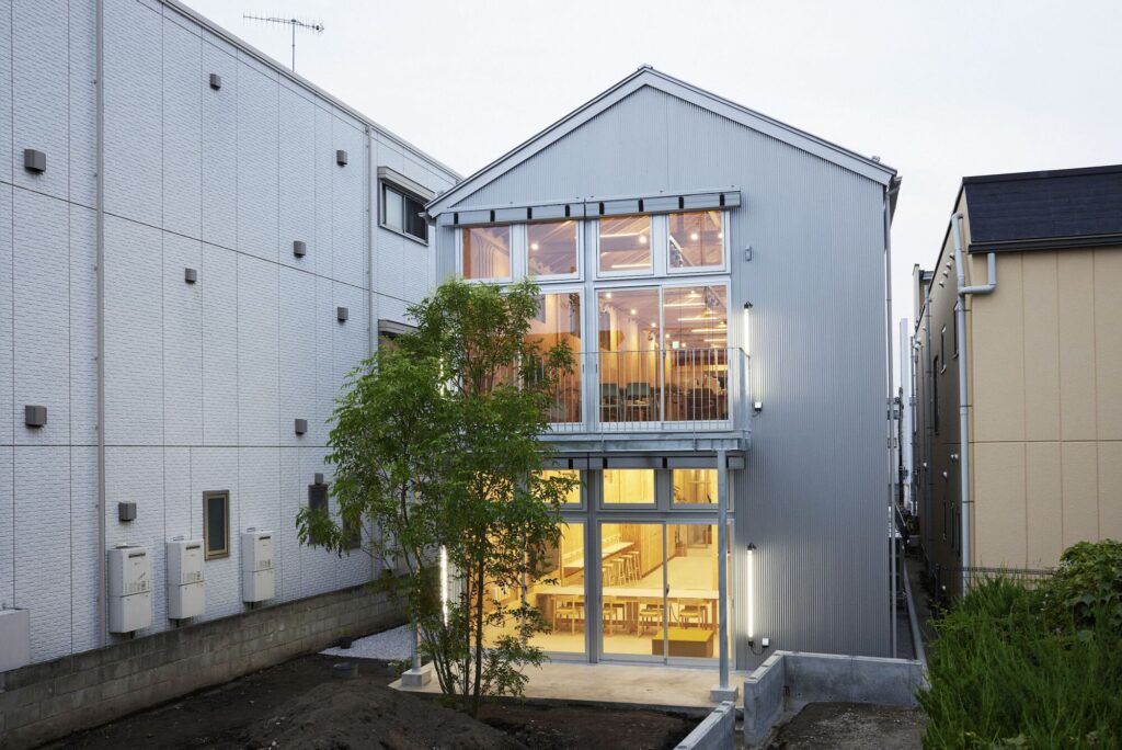 Exterior view of the Uchida Shoten headquarters featuring a silver corrugated metal facade with large glowing glass windows at dusk.