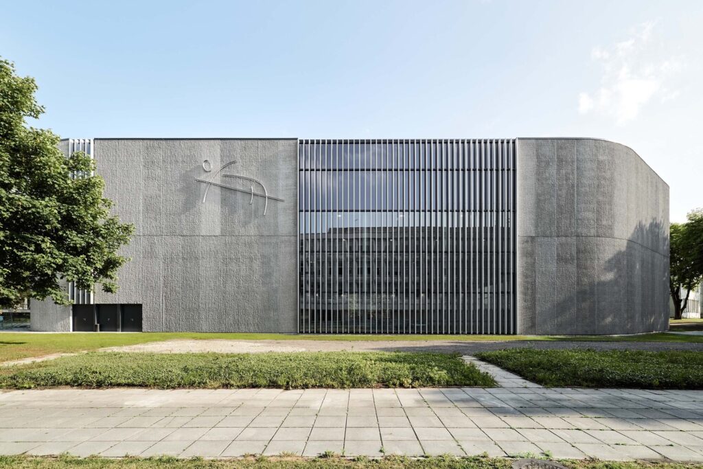 Exterior facade of the Darius and Girėnas stadium annex featuring textured oak-bark concrete and vertical glass louvers.