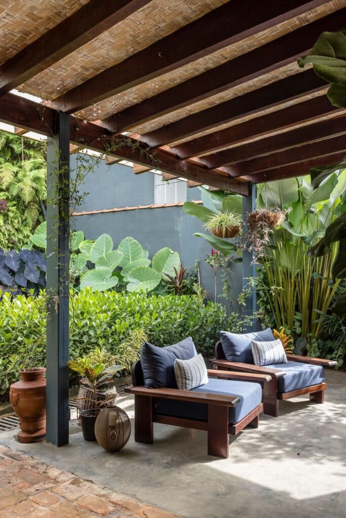 Wooden outdoor armchairs under a pergola at Heloisa and Leonardo House, surrounded by tropical plants.