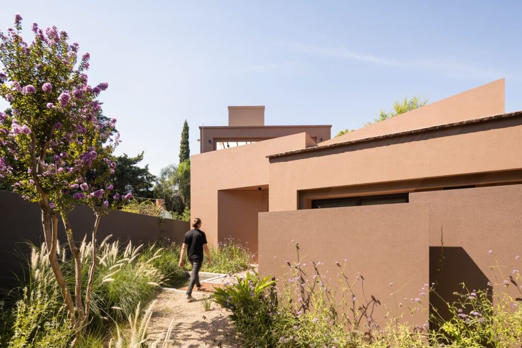 A person walking through a landscaped path toward the entrance of the earthy-toned modern residence.