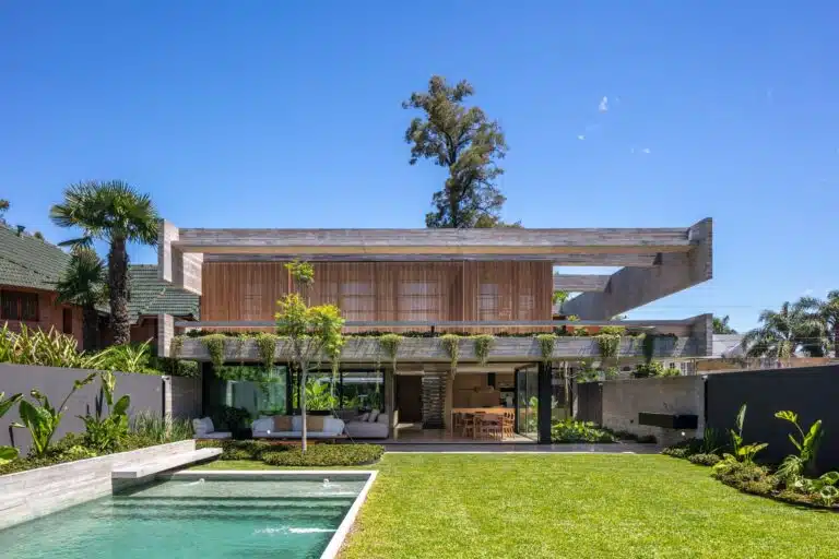 Rear view of Casa Machado by BTE Arquitectos featuring a swimming pool, green lawn, and exposed concrete structure with wooden shutters.
