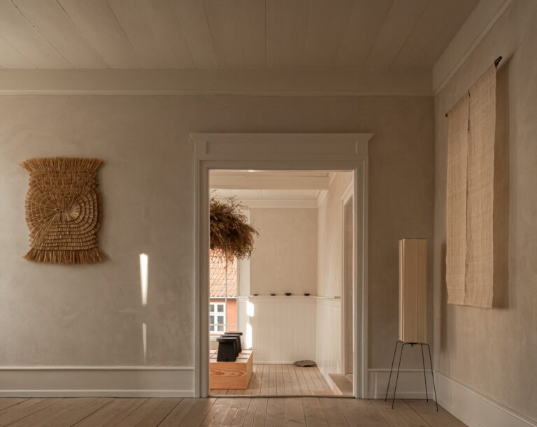 Interior gallery space with natural wood flooring, a textured woven wall hanging, and sunlight streaming through an open doorway.