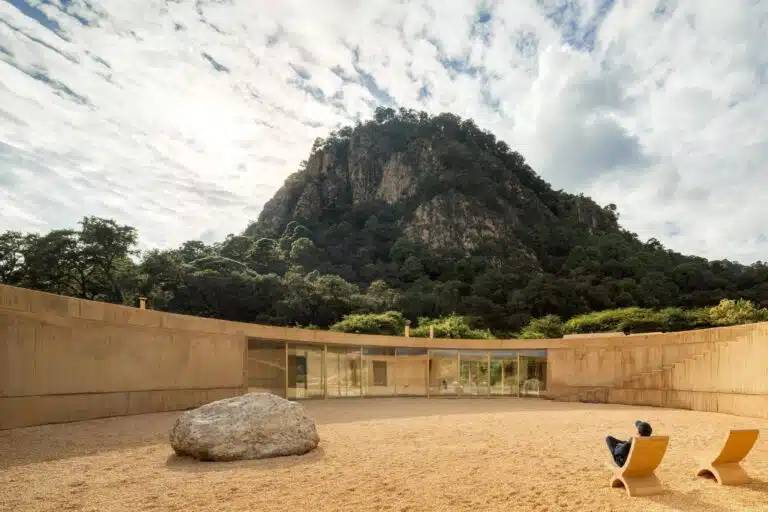 A wide view of the circular courtyard of the Degrees project with a large natural rock, a man sitting on a wooden chair, and a high mountain in the background.
