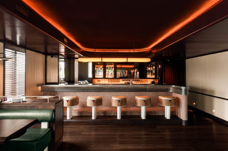 A long service bar with custom wooden stools and a metallic countertop under a glowing linear light fixture.