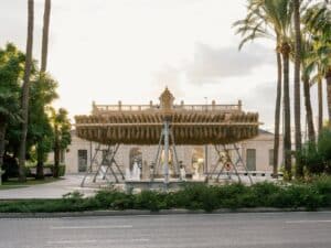 Wide shot of the Espartal pavilion featuring a suspended esparto grass structure over a public square with water fountains and palm trees.