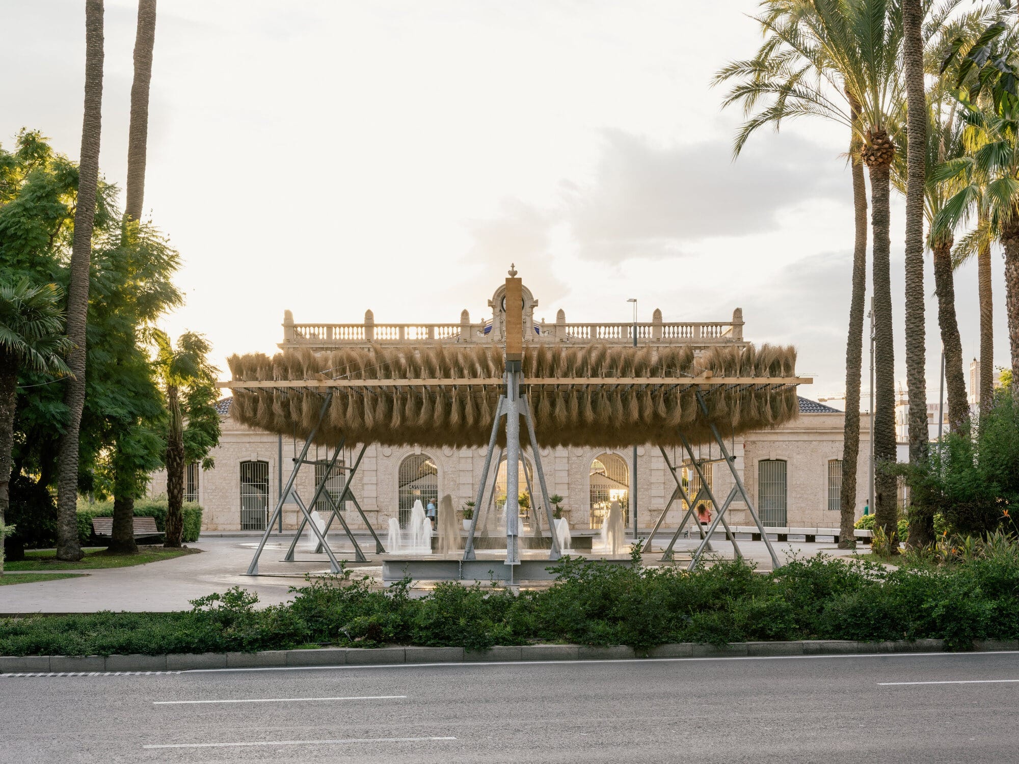 Wide shot of the Espartal pavilion featuring a suspended esparto grass structure over a public square with water fountains and palm trees.