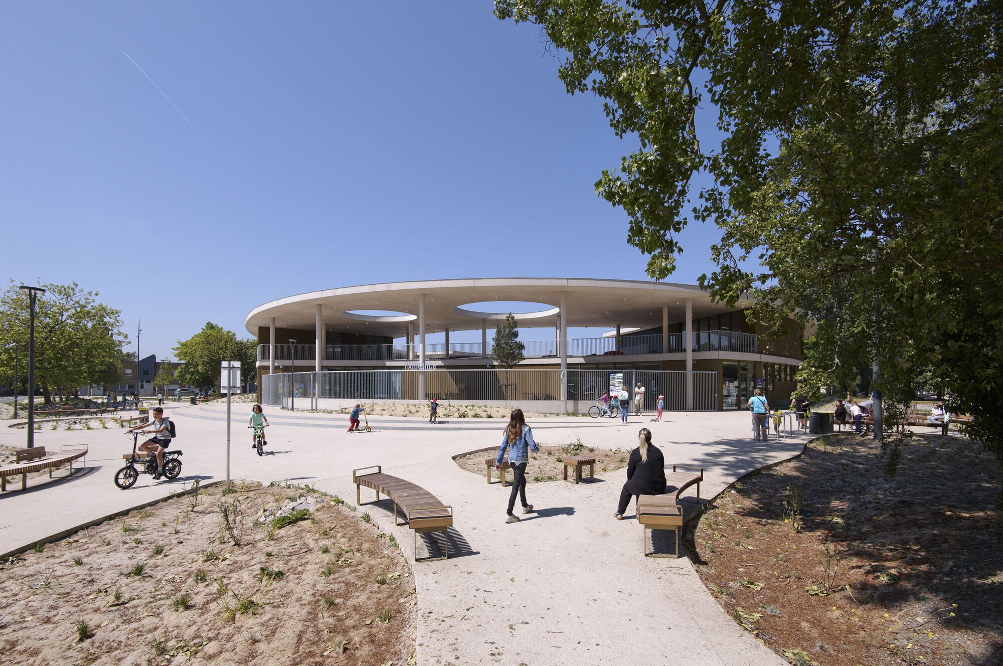 External view of L'Alliance school complex in Dunkerque showing the circular concrete canopy and public plaza with children playing.