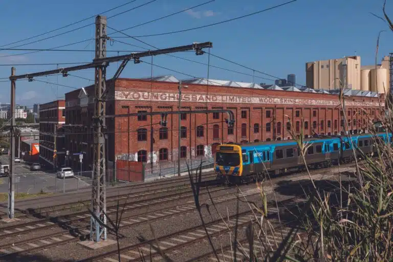 Large Victorian red brick industrial facade of the Younghusband woolstore in Melbourne with a passing Metro train and sawtooth roof silhouettes.