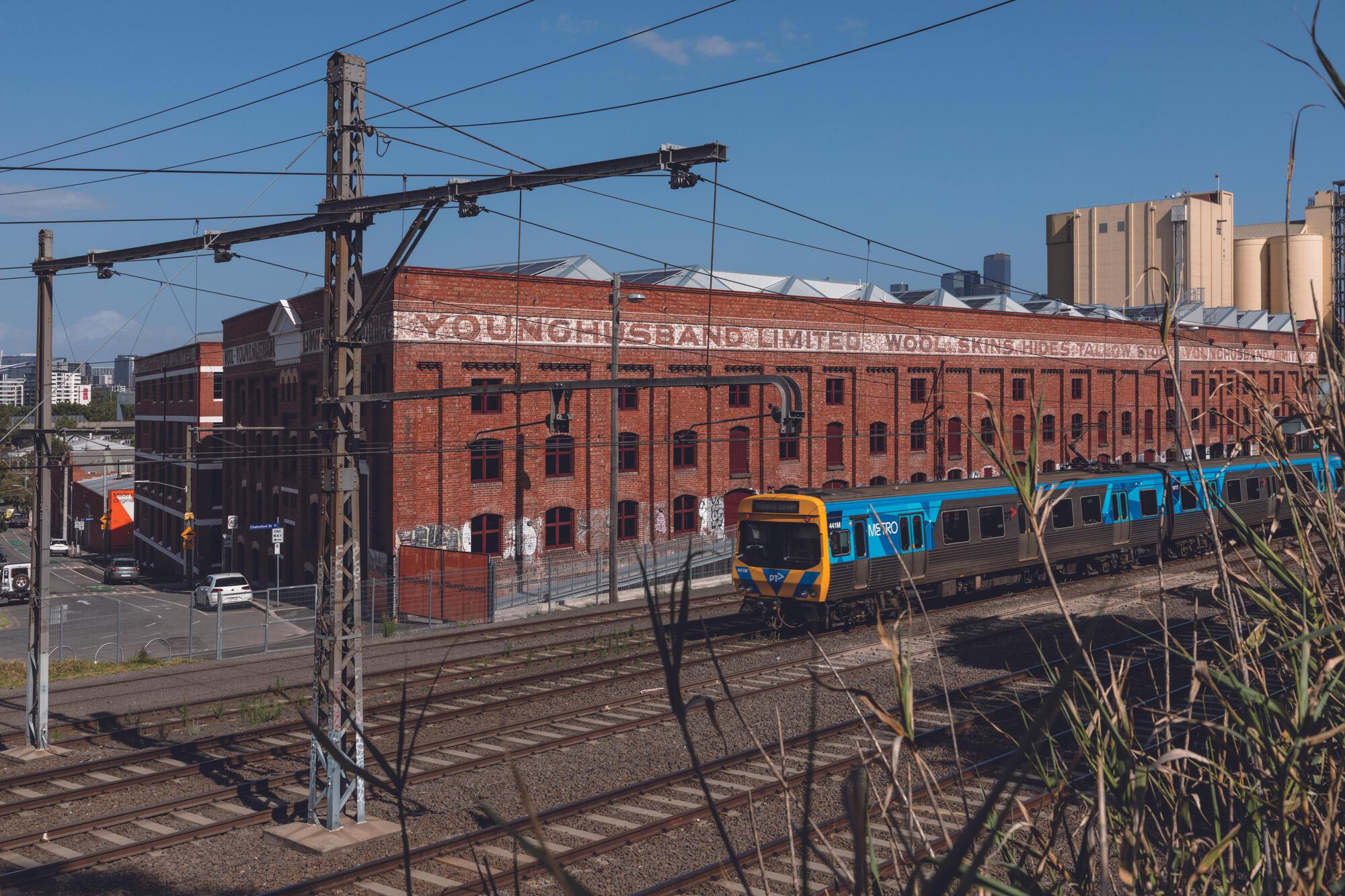Large Victorian red brick industrial facade of the Younghusband woolstore in Melbourne with a passing Metro train and sawtooth roof silhouettes.