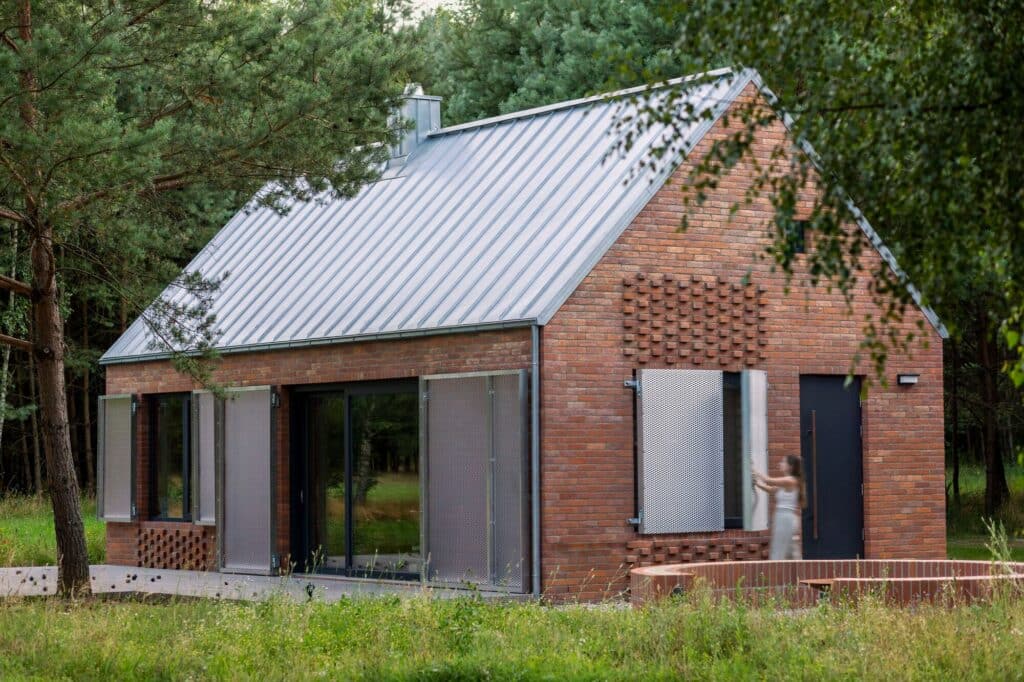 Exterior of Dom Las showing a woman opening custom metal shutters on a brick wall, surrounded by birch and pine trees.