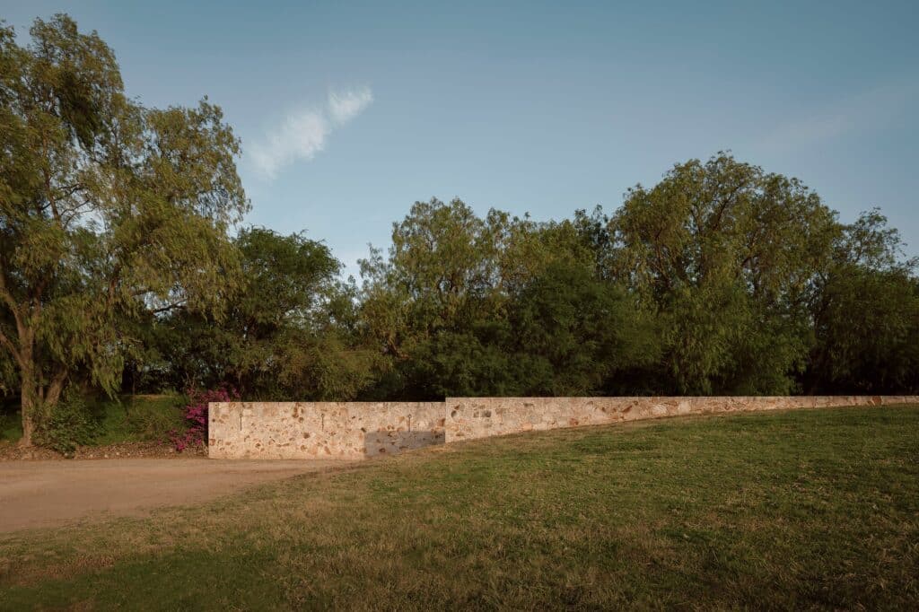 A long textured stone wall integrated into a grassy rural landscape with dense green trees under a clear blue sky.