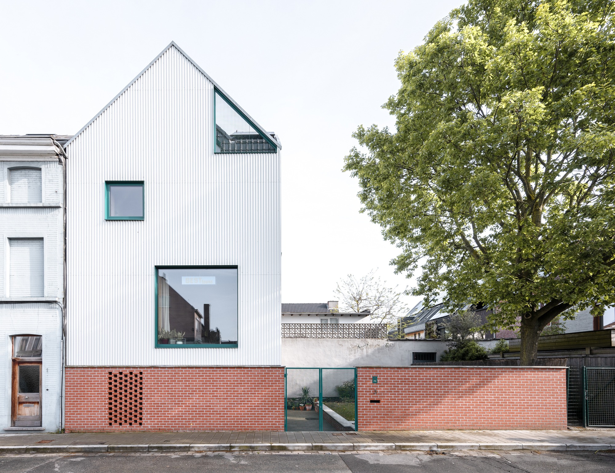Front view of Muide District House in Ghent featuring a white gable roof, green window frames, and a brick privacy wall.