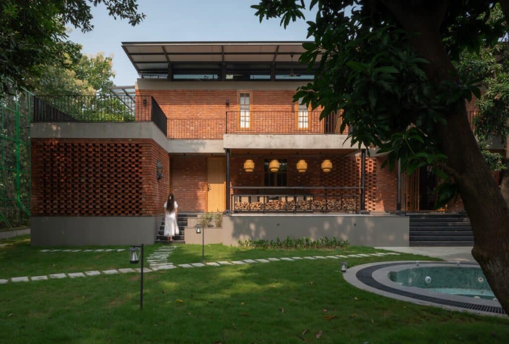 Modern farmhouse exterior with exposed red brick walls, a green lawn with stone paths, and a small circular plunge pool under the shade of trees.