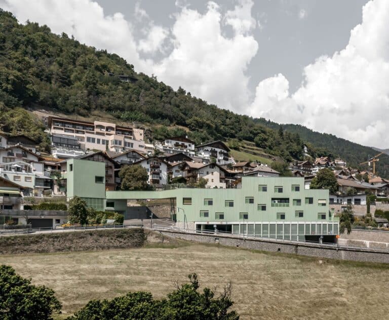 Aerial view of the Barbiano Educational Complex featuring green timber facades and a connecting bridge integrated into the South Tyrol hillside landscape.