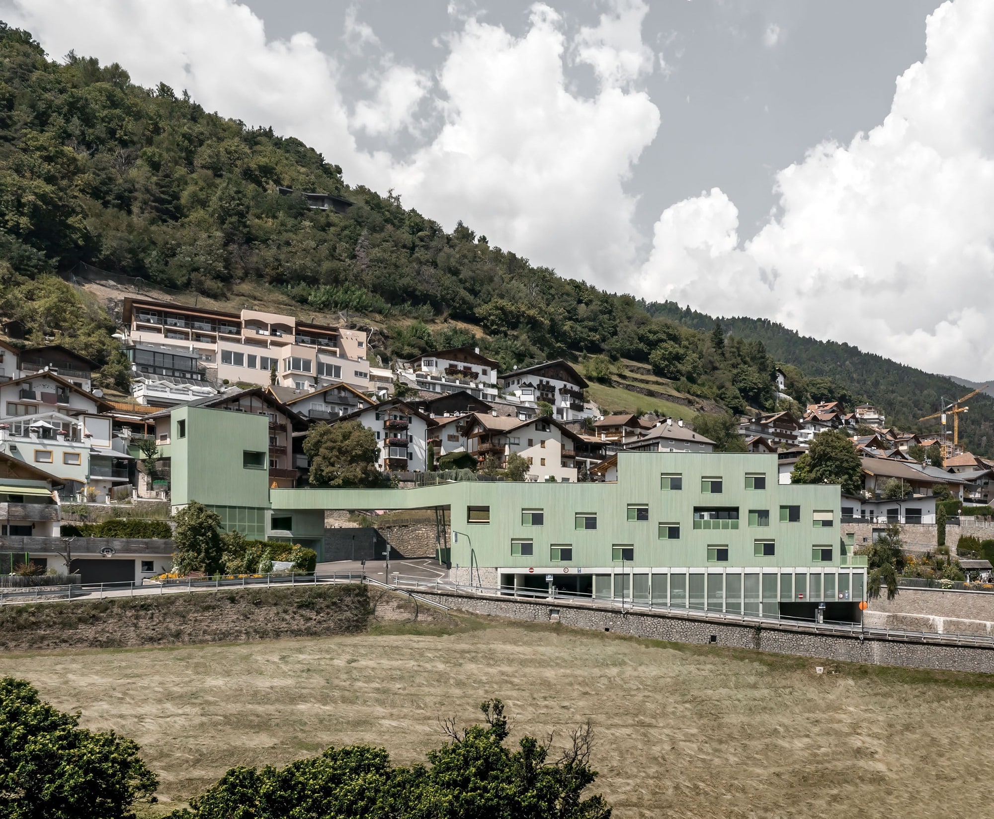Aerial view of the Barbiano Educational Complex featuring green timber facades and a connecting bridge integrated into the South Tyrol hillside landscape.