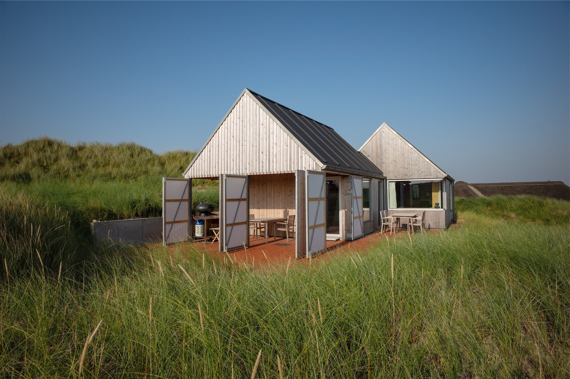 Modern wooden Kligwood Summer House with open vertical slat doors leading to an outdoor terrace surrounded by sand dunes.