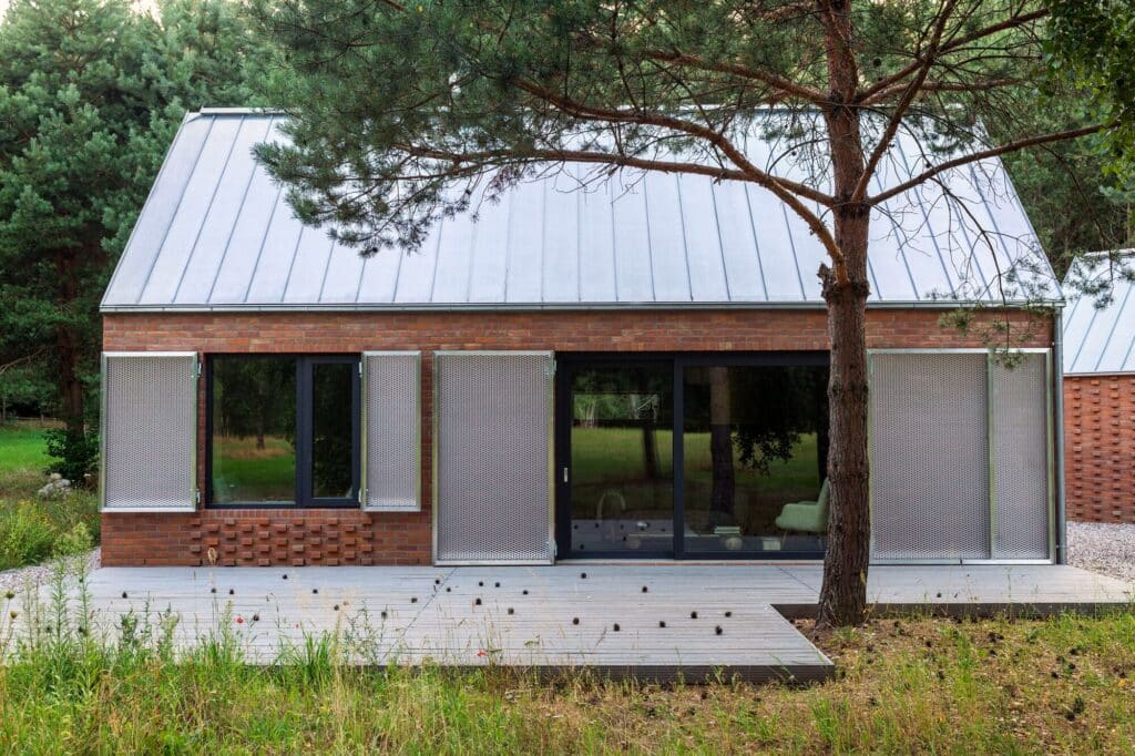 Front view of Dom Las house featuring red brick facade, metal shutters, and a wooden deck integrated with the surrounding pine forest in Poland.
