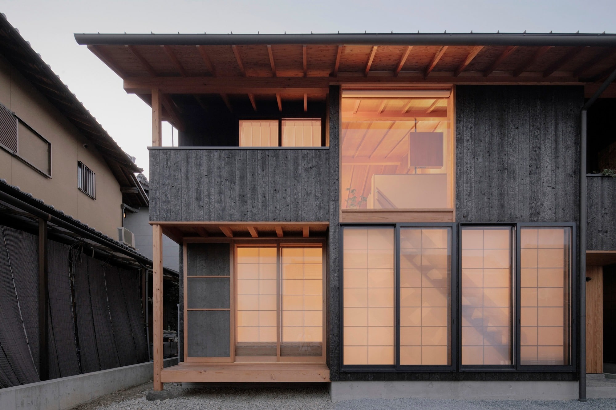 Exterior view of a modern two-story Japanese house featuring burnt cedar cladding (Shou Sugi Ban), large sliding glass doors with translucent screens, and a traditional exposed timber frame structure.