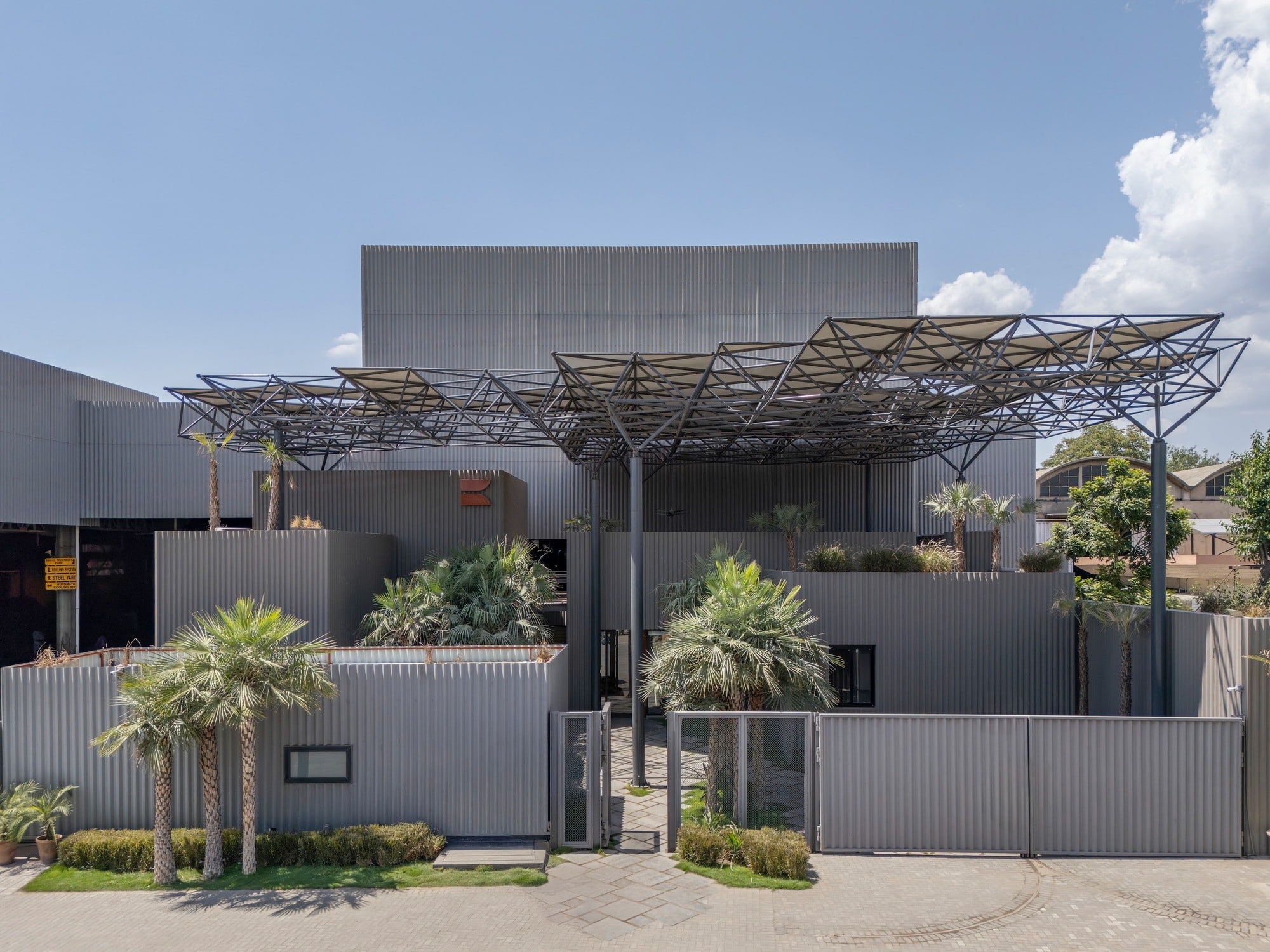 Full daylight view of Karachi Steel's entrance featuring the space frame canopy, corrugated steel cladding, and palm trees in the foreground.