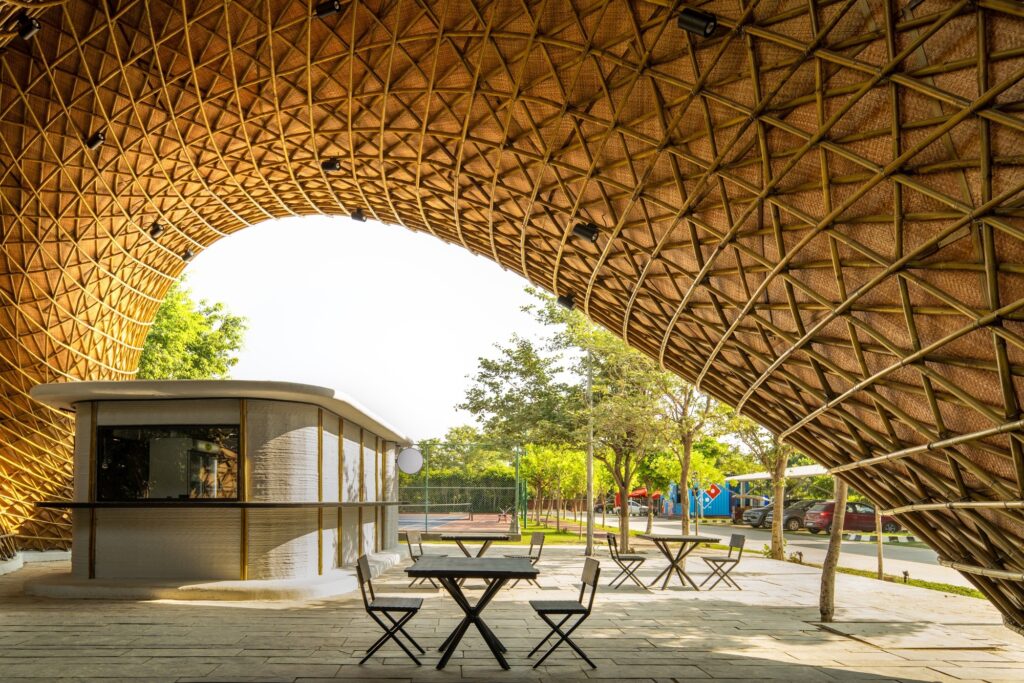 Interior-like view from under the bamboo canopy looking out towards a parking area and sports court, featuring a kiosk and seating.