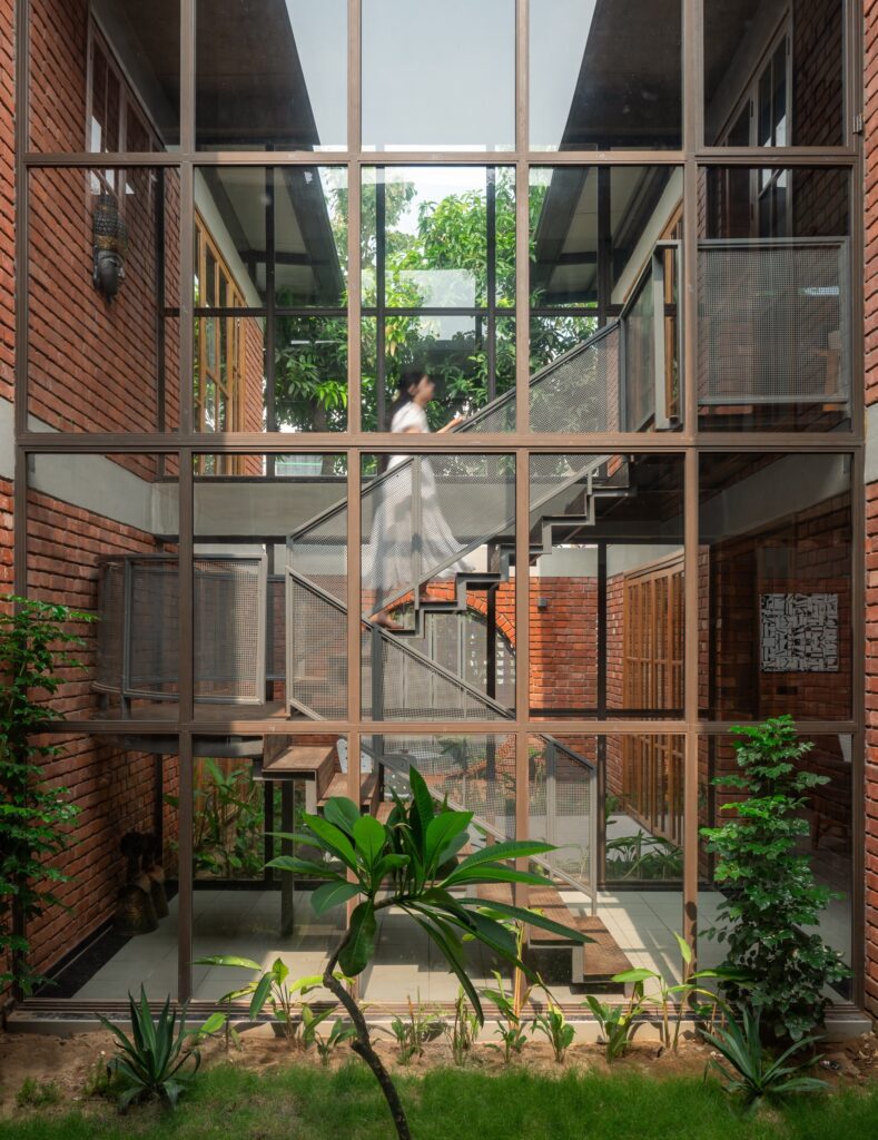 Interior metal staircase viewed through a large multi-pane glass window, flanked by exposed brick walls and indoor plants.