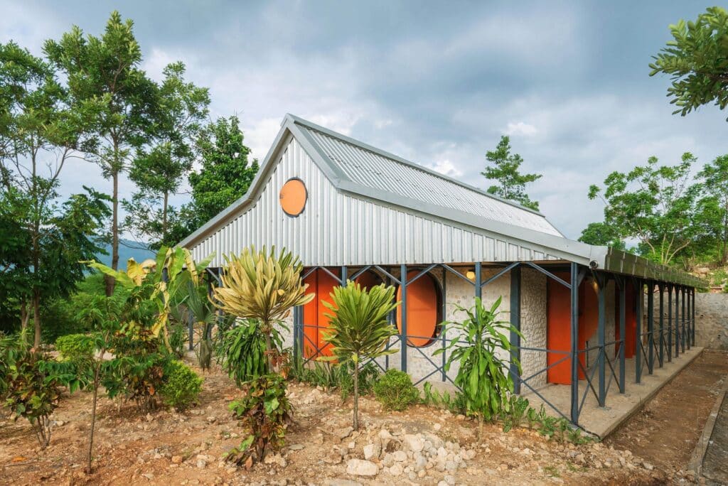 Exterior view of the community center featuring the metal roof and hybrid structure, surrounded by local flora and the jardin krey&ograve;l.