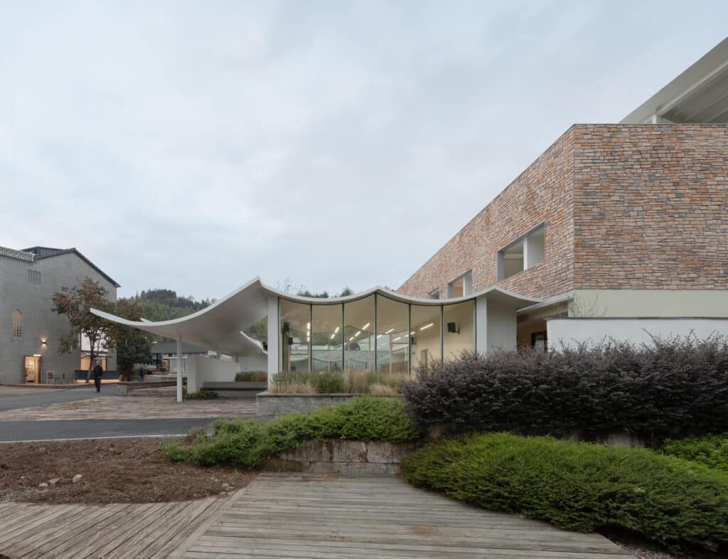 External view of the multi-functional hall with a curved white roof and glass walls at dusk.