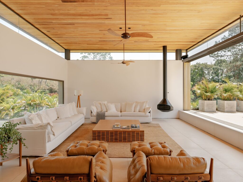 Spacious modern living room with white sofas, leather armchairs, a hanging fireplace, and floor-to-ceiling glass walls.