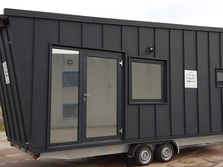 Side exterior view of the Dark Vader tiny house showing vertical matte black metal cladding and a large glass entry door on a dual-axle trailer.