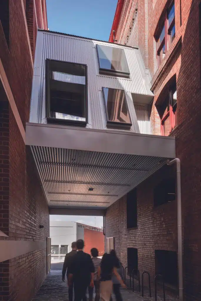 Upward view of a contemporary corrugated metal overhead bridge with large windows connecting historic brick walls at the Younghusband precinct.