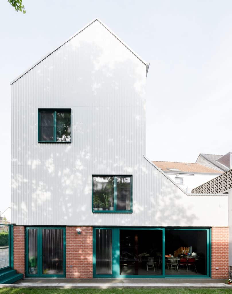 Rear facade of Muide District House showing white corrugated metal cladding, red brick base, and large glass doors opening to a green garden.