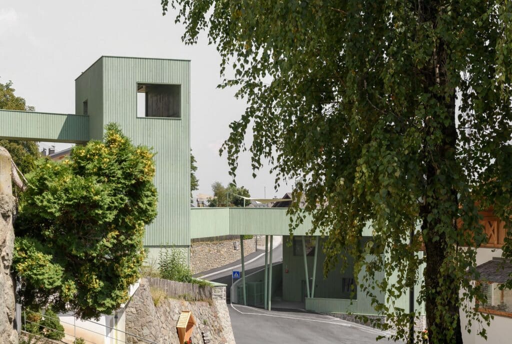 View of the green tower and bridge of the Barbiano complex framed by trees and traditional village houses.
