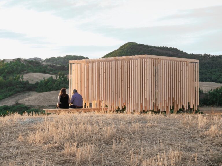 Side view of Cappella del Suono timber pavilion in Lunano Italy with two people sitting on a wooden bench overlooking rolling hills.