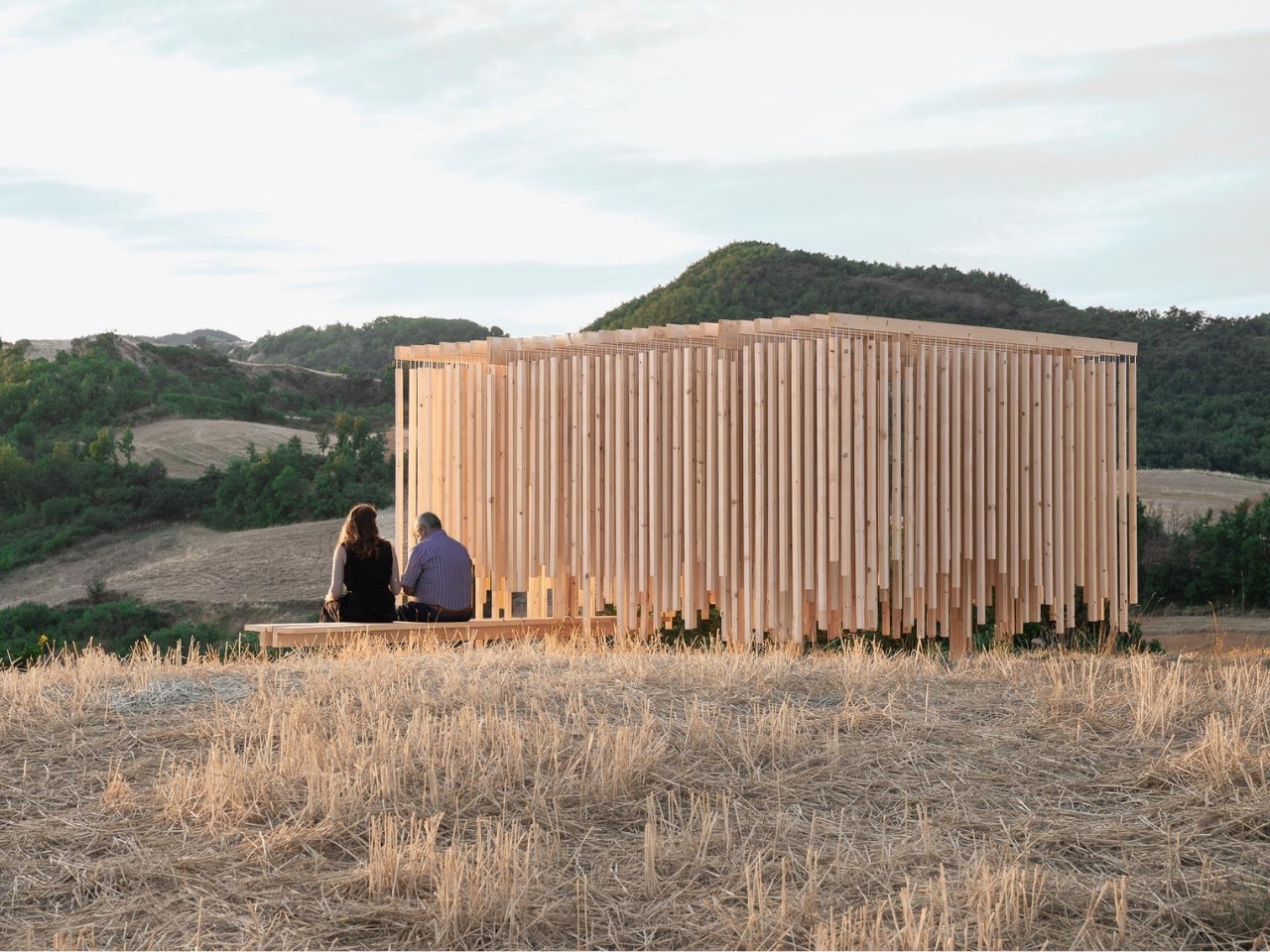 Side view of Cappella del Suono timber pavilion in Lunano Italy with two people sitting on a wooden bench overlooking rolling hills.