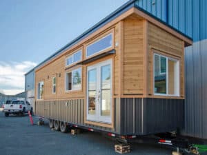 Exterior front-angle view of the Dove tiny home by Rewild Homes, featuring cedar siding, corrugated metal accents, and a triple-axle trailer.