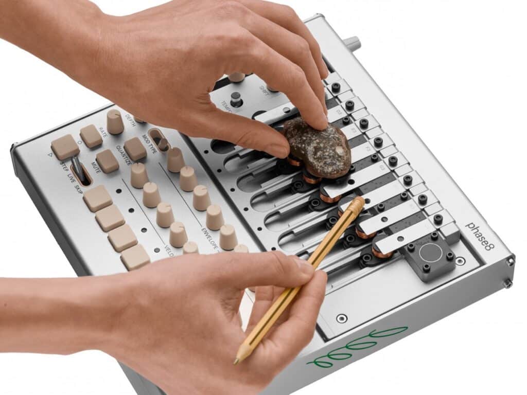 Close-up of a user&rsquo;s hands interacting with the Korg Phase8 using a natural stone and a brass stylus to strike the metal resonators, demonstrating tactile sound exploration.