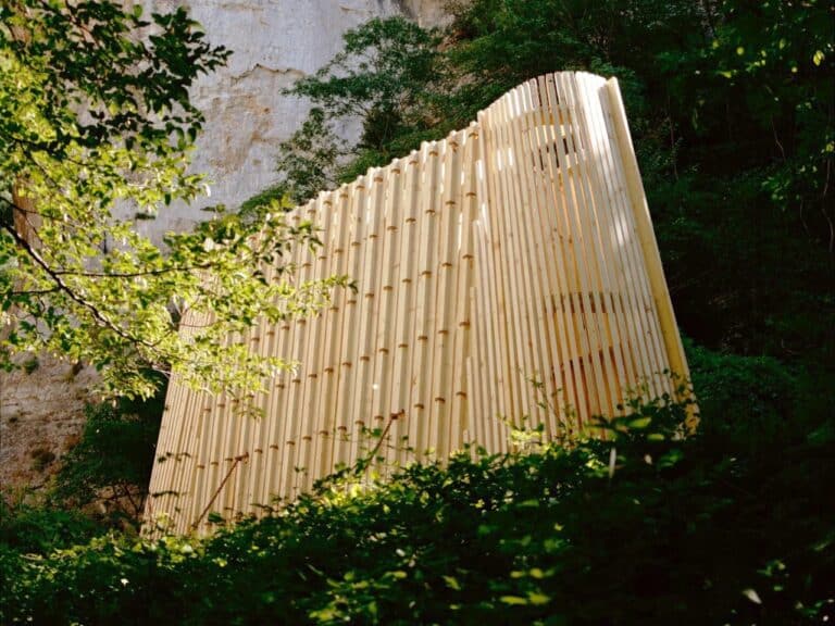 The La Barca wooden chapel illuminated by sunlight, standing tall against a massive white limestone cliff and green trees.