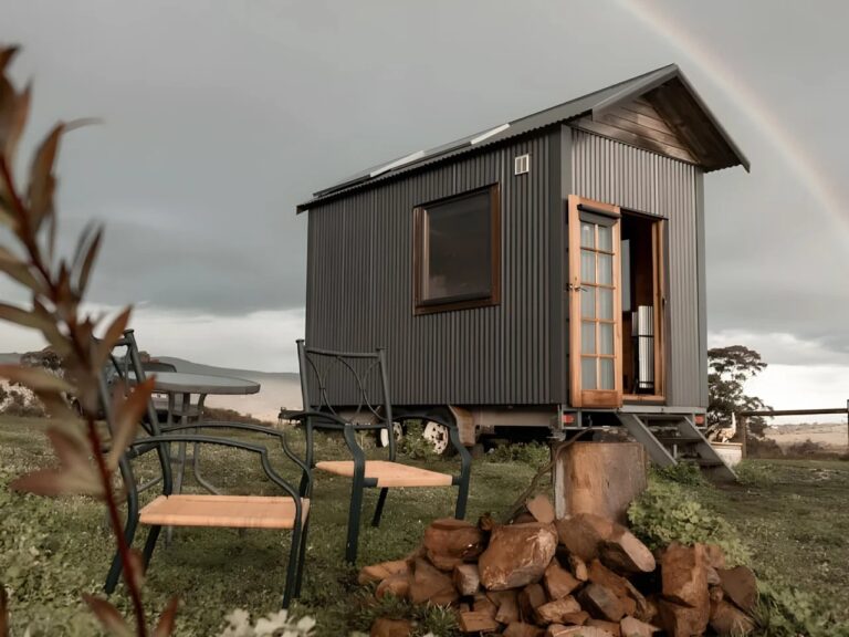 Exterior view of Tallarook Hilltop Tiny House with dark corrugated metal cladding and solar panels under a rainbow in rural Victoria.