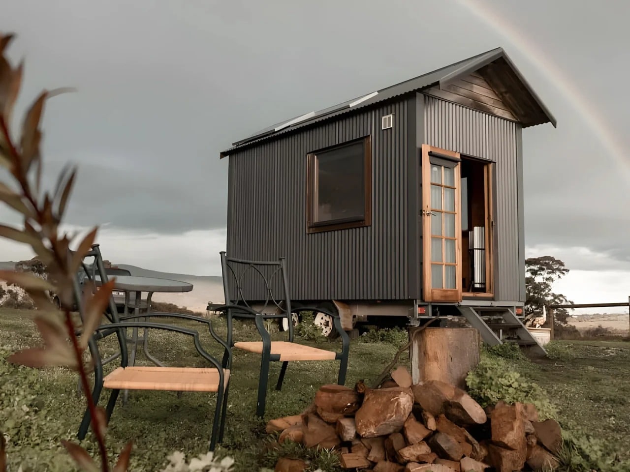 Exterior view of Tallarook Hilltop Tiny House with dark corrugated metal cladding and solar panels under a rainbow in rural Victoria.