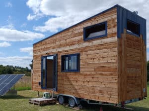 Exterior of Tiny Birdy mobile home featuring natural wood cladding, blue metal accents, and a large solar panel array in a rural setting.