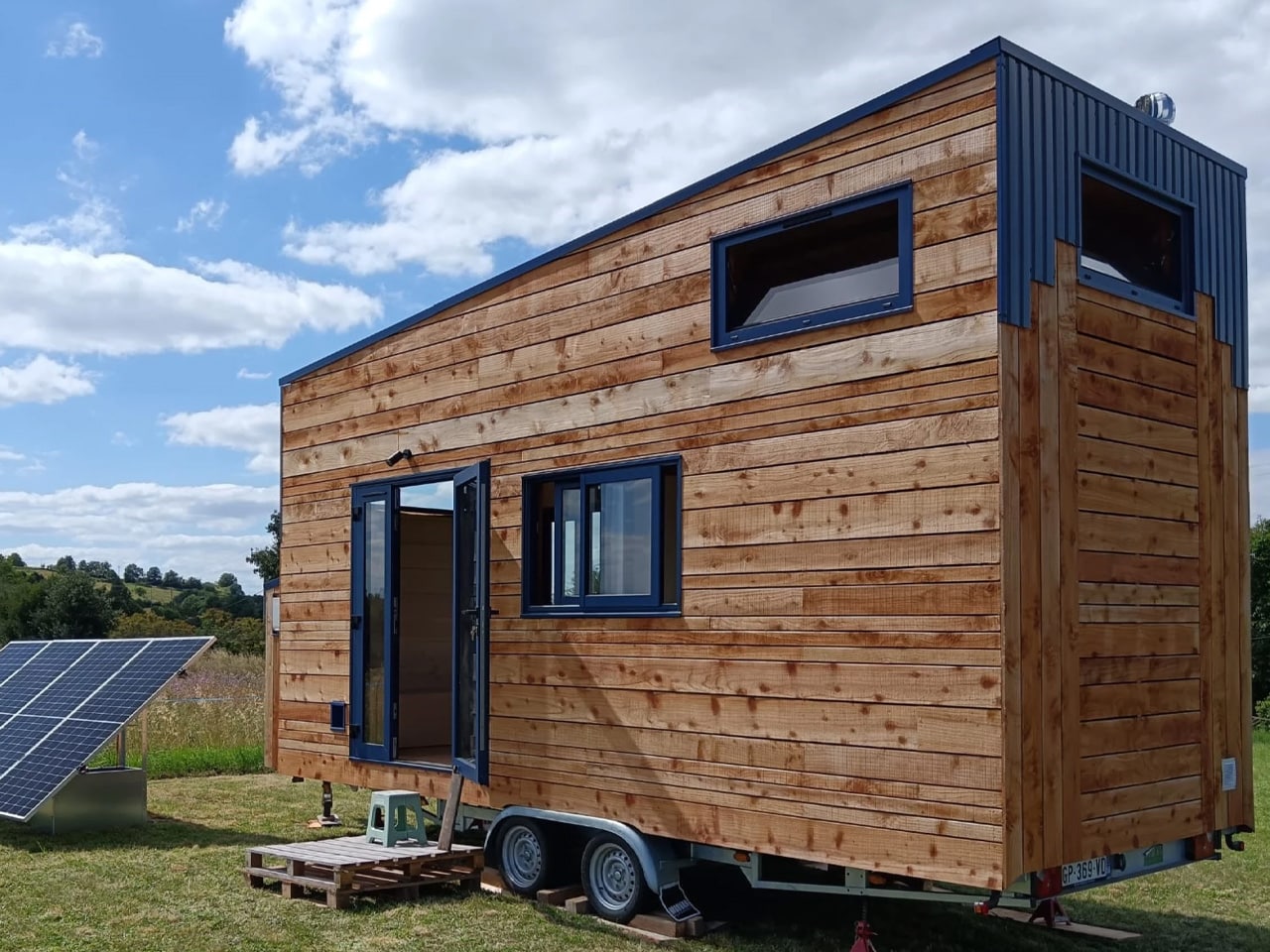 Exterior of Tiny Birdy mobile home featuring natural wood cladding, blue metal accents, and a large solar panel array in a rural setting.