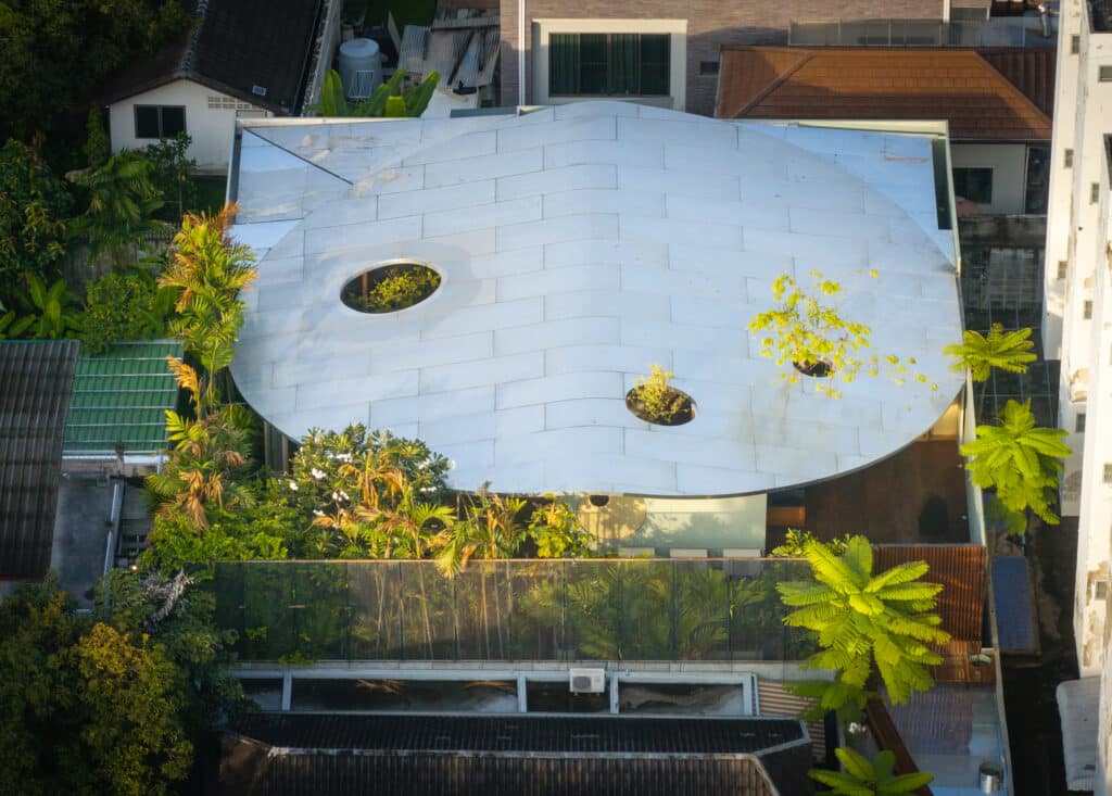 Aerial view of Terrarium House in Bangkok featuring a unique curved white roof with circular cutouts for trees, surrounded by a dense urban neighborhood.