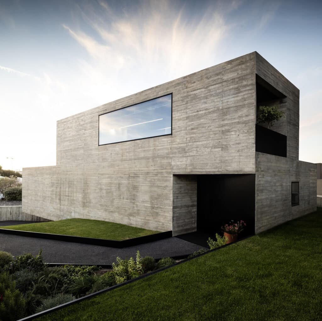 Contemporary three-story residence with large glass windows and raw concrete finish at sunset.