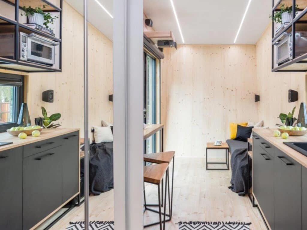 Longitudinal interior view of a tiny house featuring natural wood walls, black kitchen cabinetry, and bright linear LED ceiling lights.