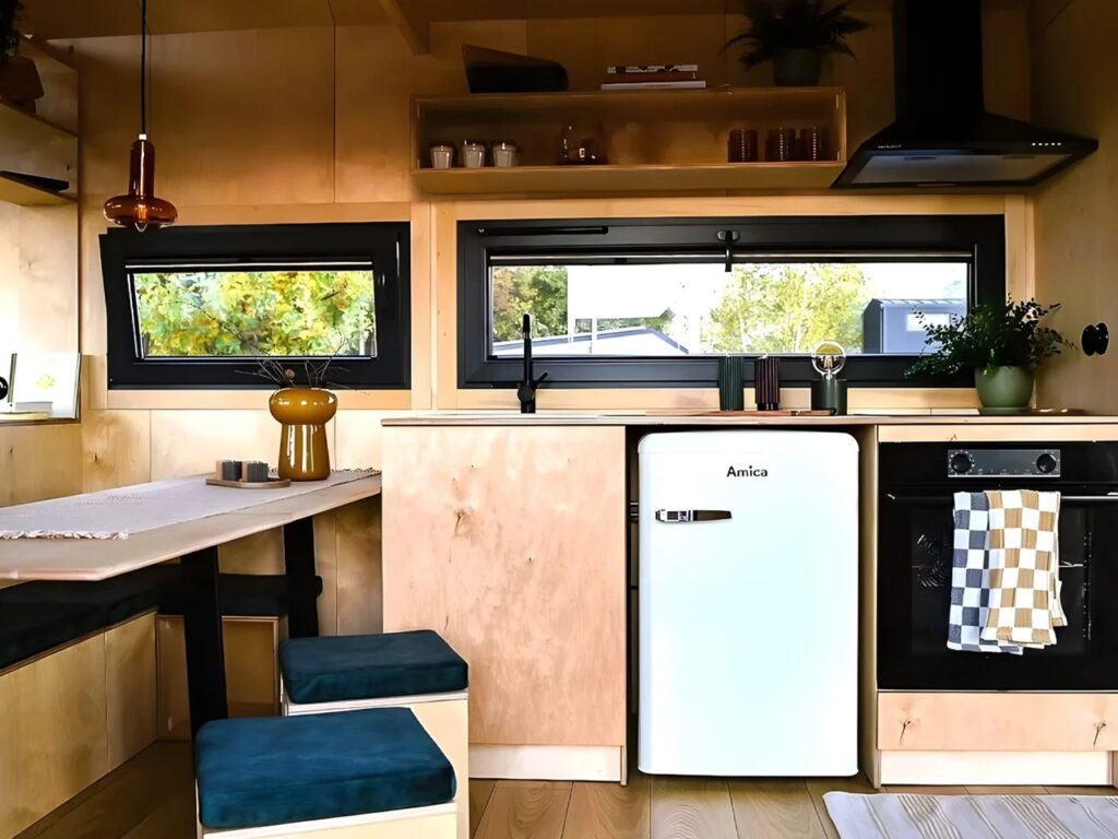 Compact modern kitchen inside Julia tiny home with plywood walls, white mini-fridge, black oven, and integrated wooden shelving.