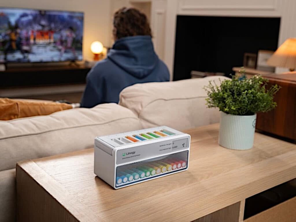Linogy battery management station placed on a wooden console in a modern living room setting with a person watching TV in the background.
